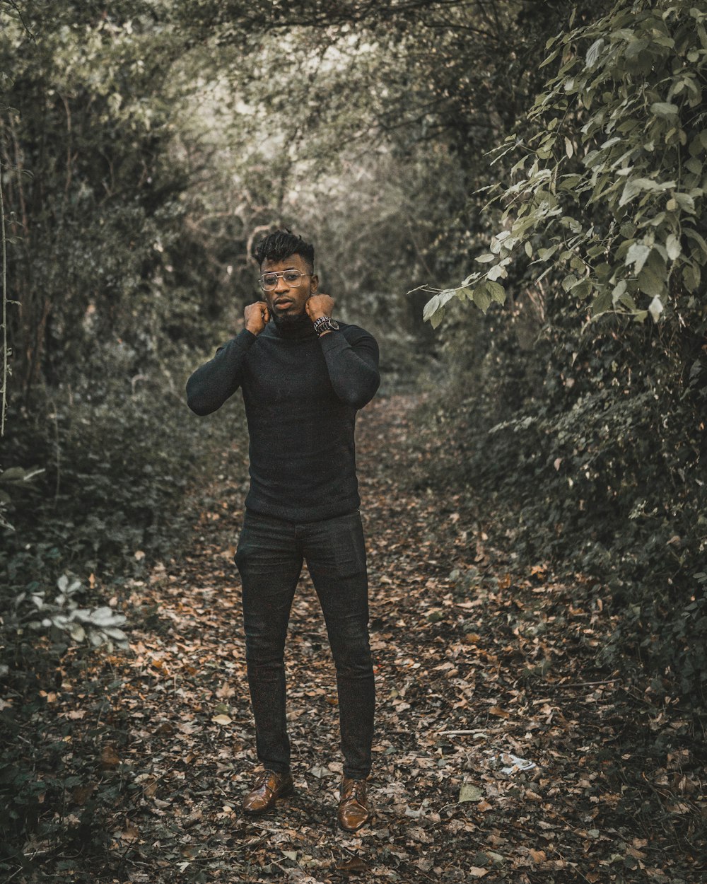 昼間、木々に囲まれた黒いタートルネックのセーターと黒いフィットパンツを着た立っている男性