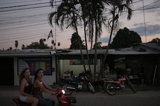 Two women are riding a red scooter on a road during dusk. There are multiple motorcycles parked in front of a building with a corrugated metal roof, which appears to be a small shop or eatery with visible tables and chairs inside. A tall palm tree is prominent in the foreground with several other trees and overhead power lines visible. There is also a flag flying in the background.