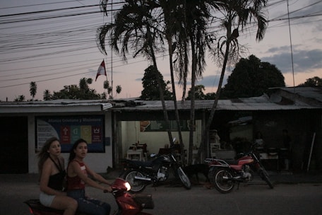 Two women are riding a red scooter on a road during dusk. There are multiple motorcycles parked in front of a building with a corrugated metal roof, which appears to be a small shop or eatery with visible tables and chairs inside. A tall palm tree is prominent in the foreground with several other trees and overhead power lines visible. There is also a flag flying in the background.