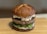 A gourmet hamburger with a glossy pretzel bun, topped with creamy sauce and thick beef patty, resting on fresh green vegetables and cucumber slices.