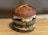 A gourmet hamburger with a glossy pretzel bun, topped with creamy sauce and thick beef patty, resting on fresh green vegetables and cucumber slices.