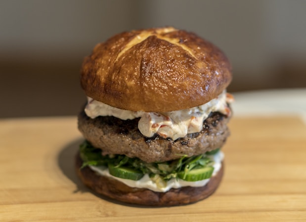 A gourmet hamburger with a glossy pretzel bun, topped with creamy sauce and thick beef patty, resting on fresh green vegetables and cucumber slices.
