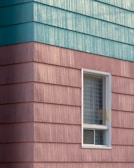 A building exterior features horizontal siding with a combination of teal and pinkish hues. The siding has a textured, weathered appearance. A single window with a white frame is visible, partially open with a grid-patterned curtain inside.
