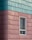 A building exterior features horizontal siding with a combination of teal and pinkish hues. The siding has a textured, weathered appearance. A single window with a white frame is visible, partially open with a grid-patterned curtain inside.