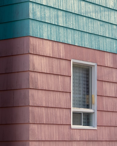 A building exterior features horizontal siding with a combination of teal and pinkish hues. The siding has a textured, weathered appearance. A single window with a white frame is visible, partially open with a grid-patterned curtain inside.