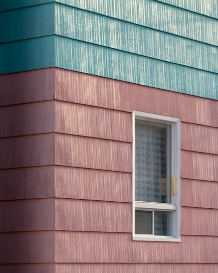 A building exterior features horizontal siding with a combination of teal and pinkish hues. The siding has a textured, weathered appearance. A single window with a white frame is visible, partially open with a grid-patterned curtain inside.