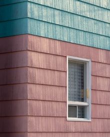 A building exterior features horizontal siding with a combination of teal and pinkish hues. The siding has a textured, weathered appearance. A single window with a white frame is visible, partially open with a grid-patterned curtain inside.