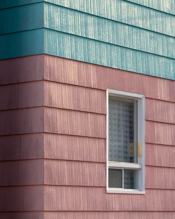 A building exterior features horizontal siding with a combination of teal and pinkish hues. The siding has a textured, weathered appearance. A single window with a white frame is visible, partially open with a grid-patterned curtain inside.
