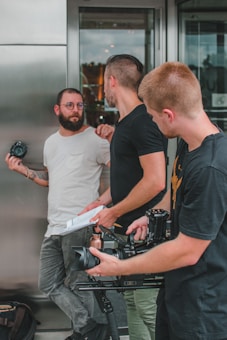 Three men are standing outside, engaged in conversation. One is holding a camera, another has a notebook, and the third is managing a video camera mounted on a stabilizer. They appear to be involved in a filmmaking or photography activity.
