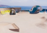 A skate park with smooth concrete surfaces features various ramps and an arched passageway. Graffiti decorates one of the walls, with yellow and grey tags. The park overlooks a scenic ocean view, with clear blue water stretching to the horizon under a cloudy sky.
