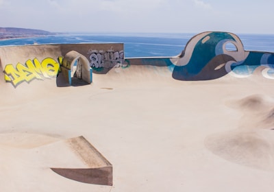 A skate park with smooth concrete surfaces features various ramps and an arched passageway. Graffiti decorates one of the walls, with yellow and grey tags. The park overlooks a scenic ocean view, with clear blue water stretching to the horizon under a cloudy sky.