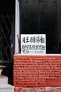 A sign with Chinese characters is placed in the window of a building with a red brick facade. The top portion of the facade is a window with a black interior. There is a noticeable iron gate to the left, partially open, revealing some stairs going up.