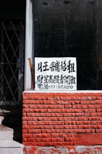 A sign with Chinese characters is placed in the window of a building with a red brick facade. The top portion of the facade is a window with a black interior. There is a noticeable iron gate to the left, partially open, revealing some stairs going up.