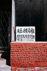 A sign with Chinese characters is placed in the window of a building with a red brick facade. The top portion of the facade is a window with a black interior. There is a noticeable iron gate to the left, partially open, revealing some stairs going up.