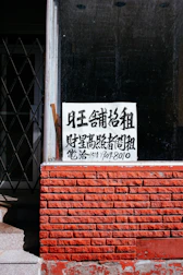 A sign with Chinese characters is placed in the window of a building with a red brick facade. The top portion of the facade is a window with a black interior. There is a noticeable iron gate to the left, partially open, revealing some stairs going up.