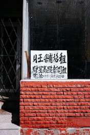 A sign with Chinese characters is placed in the window of a building with a red brick facade. The top portion of the facade is a window with a black interior. There is a noticeable iron gate to the left, partially open, revealing some stairs going up.