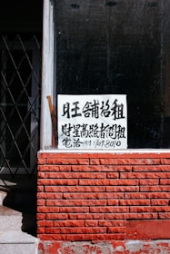 A sign with Chinese characters is placed in the window of a building with a red brick facade. The top portion of the facade is a window with a black interior. There is a noticeable iron gate to the left, partially open, revealing some stairs going up.