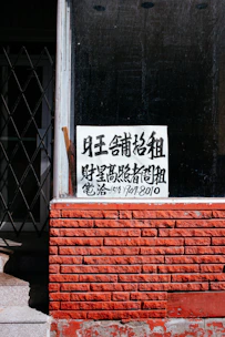 A sign with Chinese characters is placed in the window of a building with a red brick facade. The top portion of the facade is a window with a black interior. There is a noticeable iron gate to the left, partially open, revealing some stairs going up.
