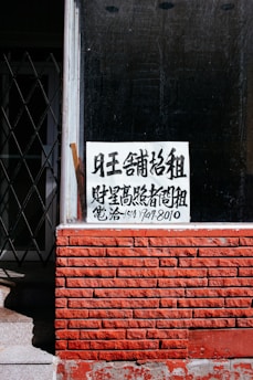 A sign with Chinese characters is placed in the window of a building with a red brick facade. The top portion of the facade is a window with a black interior. There is a noticeable iron gate to the left, partially open, revealing some stairs going up.
