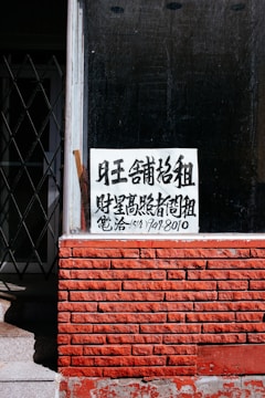 A sign with Chinese characters is placed in the window of a building with a red brick facade. The top portion of the facade is a window with a black interior. There is a noticeable iron gate to the left, partially open, revealing some stairs going up.
