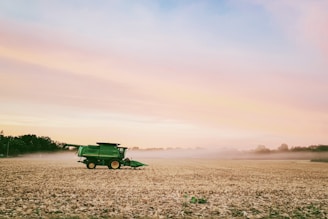 A green combine harvester working in a large, open agricultural field with harvested crops. The sky is filled with gentle hues of orange, pink, and blue, creating a serene sunset scene. Trees line the horizon in the background.