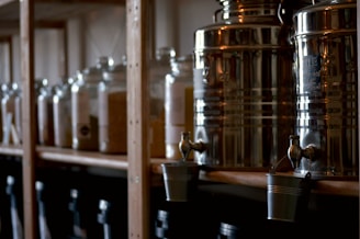 A set of metal dispensers with taps, likely for liquid storage, are lined up on a wooden shelf. Below them, small metallic buckets hang from the taps. The background features large transparent jars, possibly holding grains or powders, also placed on the shelf.