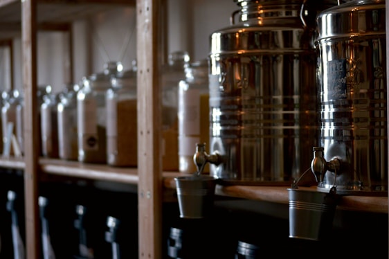 A set of metal dispensers with taps, likely for liquid storage, are lined up on a wooden shelf. Below them, small metallic buckets hang from the taps. The background features large transparent jars, possibly holding grains or powders, also placed on the shelf.
