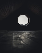 A dimly lit attic room features a wooden floor with visible grains and textures. The focal point is an octagonal window that allows a soft light to filter through, partially illuminating the space. Shadows dominate the upper corners, creating a mysterious and secluded atmosphere.