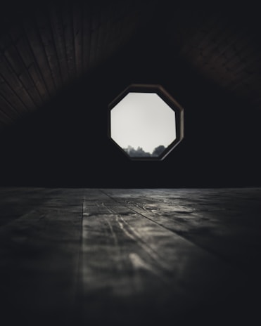 A dimly lit attic room features a wooden floor with visible grains and textures. The focal point is an octagonal window that allows a soft light to filter through, partially illuminating the space. Shadows dominate the upper corners, creating a mysterious and secluded atmosphere.
