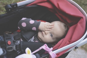 A baby is lying in a stroller with a red canopy. The baby is wearing a polka dot sleeve and appears to be playing by covering one eye with a hand. The stroller has black and red straps and a pink clip.