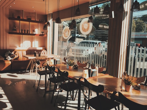 The interior of a cozy coffee shop is illuminated by warm sunlight streaming through large windows. There are wooden tables and chairs arranged neatly, and potted plants add a touch of greenery to the space. A comfortable brown leather couch is situated against the wall, and shelves with small decorative items hang above it. Outside, a few cars are parked and a person is passing by.