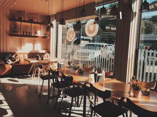 The interior of a cozy coffee shop is illuminated by warm sunlight streaming through large windows. There are wooden tables and chairs arranged neatly, and potted plants add a touch of greenery to the space. A comfortable brown leather couch is situated against the wall, and shelves with small decorative items hang above it. Outside, a few cars are parked and a person is passing by.