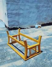 An upside-down yellow wooden table is placed on a rough, blue-textured surface in an outdoor space. In the background, there is a gray, weathered wall, beside which are two containers, one appearing to be a bottle and the other a small bowl.