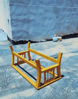 An upside-down yellow wooden table is placed on a rough, blue-textured surface in an outdoor space. In the background, there is a gray, weathered wall, beside which are two containers, one appearing to be a bottle and the other a small bowl.