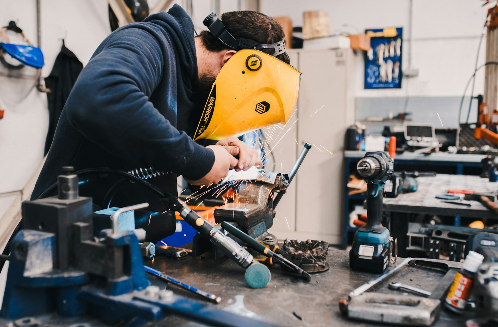 A person in a navy hoodie holds a yellow welding mask to their face while welding a small steel assembly clamped in a vise on a cluttered workbench.