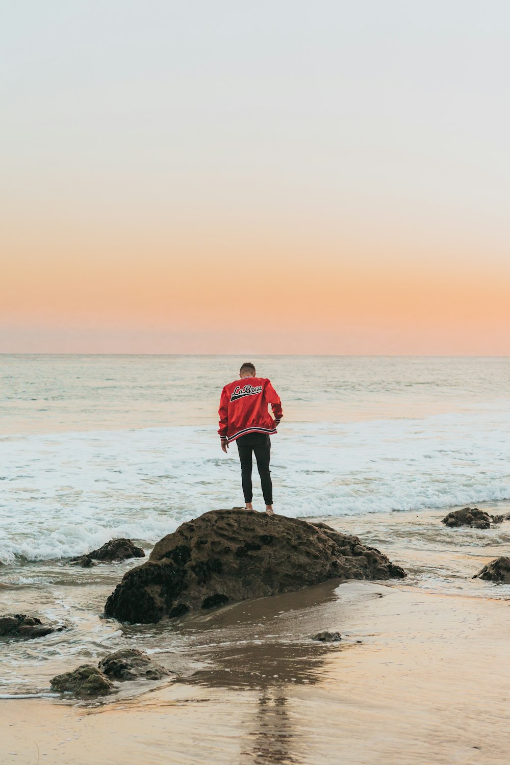 Mann in orangefarbener Jacke, der auf einem Küstenfelsen in der Nähe des Strandes steht