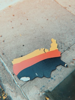 A flat, wooden cutout of the United States is placed on a concrete sidewalk. The cutout is painted in three horizontal stripes, with yellow on top, red in the middle, and black at the bottom.