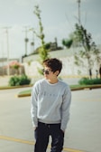A person standing outdoors, wearing a light grey sweatshirt with text and dark pants. They are wearing sunglasses and have short, tousled hair. The background includes a street with painted lines, greenery, and blurred utility structures under a clear sky.
