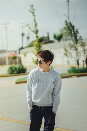 A person standing outdoors, wearing a light grey sweatshirt with text and dark pants. They are wearing sunglasses and have short, tousled hair. The background includes a street with painted lines, greenery, and blurred utility structures under a clear sky.