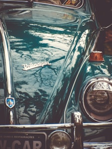 A vintage Volkswagen car with a shiny teal exterior reflecting a pattern of tree branches. The car features a classic headlight and a distinctive emblem, with a portion of a license plate visible at the bottom. An orange indicator light is located next to the headlight.