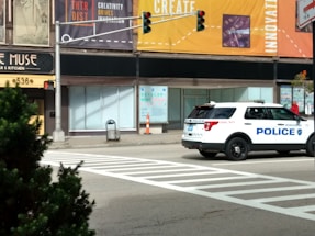A police SUV is stopped at an intersection on a city street. There are noticeable storefronts and posters advertising creativity and innovation. A pedestrian crossing is painted on the road, and a traffic light is visible. In the foreground, there is greenery on a sidewalk.