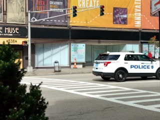 A police SUV is stopped at an intersection on a city street. There are noticeable storefronts and posters advertising creativity and innovation. A pedestrian crossing is painted on the road, and a traffic light is visible. In the foreground, there is greenery on a sidewalk.