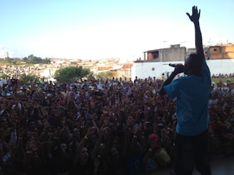A performer stands on stage with a microphone, hand raised, facing a large, enthusiastic crowd. The audience is packed closely together, with many individuals raising their hands in the air. The background includes urban buildings and a clear sky.