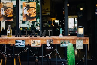 A wooden countertop adorned with various decorative items including a metal 'MIAMI' sign, a small model of the Statue of Liberty, a black candle lantern, and a cactus. Below the countertop, there are several retro photographs and postcards strung along, and in the background, there is a view into a restaurant with posters on the glass doors and windows.