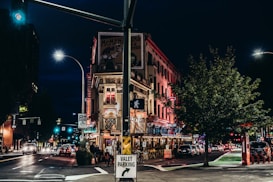 A bustling urban street corner at night featuring a historic looking red-brick building adorned with neon lights and signage. Cars drive by under illuminated streetlights, and a restaurant or bar beneath the well-lit building has outdoor seating. A valet parking sign is visible in the foreground.