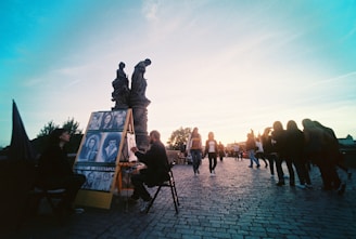 A street artist is seated, drawing portraits on an easel as people walk by in the background. The scene is set on a cobblestone path, likely on or near a bridge with historical statues elevated in the background. The sun casts a warm glow, creating a serene and lively atmosphere. Nearby, a person observes the artist's work.