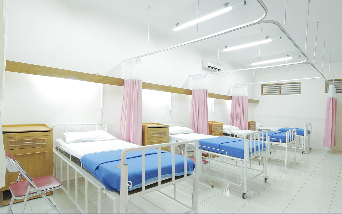 a row of 4 hospital beds each separated by pink curtains.  There is a light brown dresser next to each bed and the room is lit by flourescent lights.