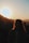 A silhouette of a person with curly hair standing against the backdrop of a setting sun, with mountains and trees in the distance creating a serene landscape.