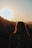 A silhouette of a person with curly hair standing against the backdrop of a setting sun, with mountains and trees in the distance creating a serene landscape.