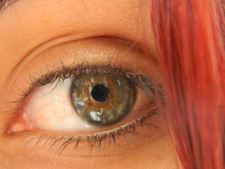 A close-up view of a human eye with detailed features such as the iris, pupil, and eyelashes. The iris is a mix of green and brown colors, and there is a strand of red hair partially obscuring part of the face.